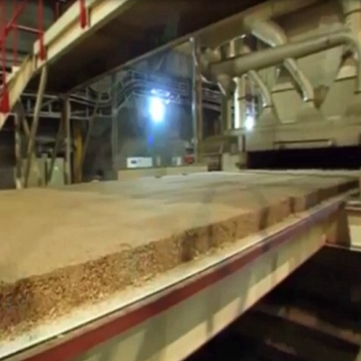 A mat of wood chips and resin moving on a conveyor toward a heated press